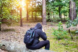 A man with a backpack in a hood sits on a fallen tree in the middle of a clearing in the forest. Survival in the wild. Dehydration of the body. Mushroom picker lost in the forest. photo