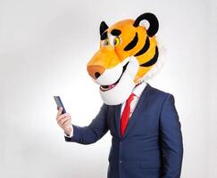 A man in a business suit and a tiger mask on his head looks into a smartphone on a white background, humor. photo