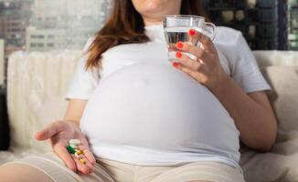 A pregnant girl holds a cup of water and a handful of pills in her palm. Painkillers and vitamins during pregnancy. photo