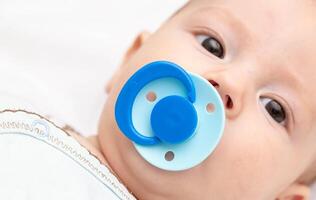 A 5 month old baby with a blue pacifier in his mouth looks at the camera. The concept of the habit of sucking on the nipple, the sucking reflex in babies. Copy space for text photo