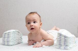 A little boy at the age of 5 months lies in a diaper on a white background and looks at the camera. Stack of diapers, anti-leak, soft and comfortable. Copy space for text photo