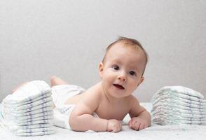 A little boy at the age of 5 months lies in a diaper on a white background and looks at the camera. Stack of diapers, anti-leak, soft and comfortable. Copy space for text photo