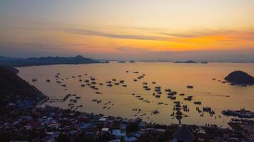 Aerial view of Labuan Bajo harbor at sunset, showcasing numerous pinisi boats docked, with charming local residences surrounding the vibrant scene. photo