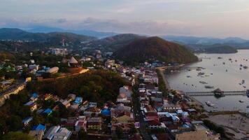 Aerial view of Labuan Bajo harbor at sunset, showcasing numerous pinisi boats docked, with charming local residences surrounding the vibrant scene. photo