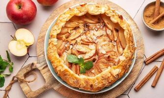Homemade apple tart with almond toppings on a wooden board surrounded by fresh apples and cinnamon photo