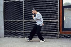 A man wearing a gray sweater and black pants dances in front of a chain-link fence in the daytime photo
