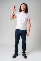 A young man in a white t-shirt and jeans confidently shows an okay hand gesture photo