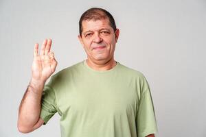 A man with a slight smile making an OK hand gesture against a neutral background photo