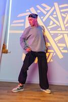 A young man wearing a bandana and a grey sweatshirt strikes a pose in a dance studio with a colorful backdrop photo