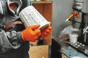 Worker in protective gear holds a paint container, preparing for spray painting in a workshop during the day photo