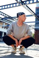A young man in a grey sweater and black pants sits in a squat position on a paved surface under a metal structure photo