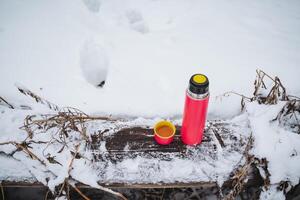 A thermos with a mug stands on a wooden table in the forest in winter, pink tourist utensils, thermo flask. photo