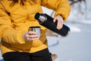 Woman in yellow jacket pouring coffee from thermos wearing gloves photo