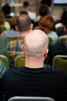 A bald man can be seen sitting in a classroom surrounded by other people photo