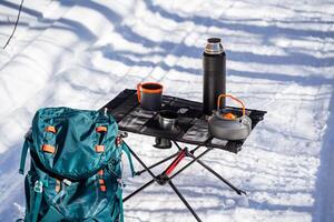 Hiking equipment for hiking stands on the snow in winter in the forest, folding table made of mesh and aluminum legs. Tableware for food photo