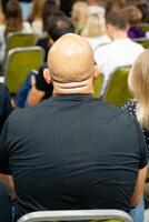 A bald man is sitting comfortably in a chair before a crowd of people photo