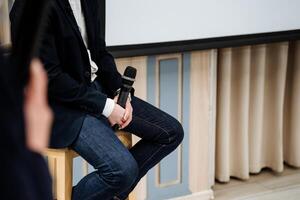 A man is sitting on a bar stool and holding a black microphone in his hands, a coach is giving a speech at a conference, a guy is giving a speech on stage. photo