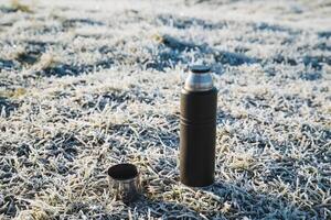 Thermos standing on the ground in cold weather, tea mug on a hike, camping utensils, hot drink in a metal flask. photo