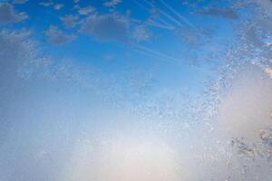 Frost draws snowflakes on the glass, frozen window in cold winter, blue sky shines through the glass. photo