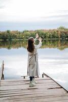 A girl standing in a raincoat on a pyron, a dance of a young beauty in a coat on the bridge, a rear view of a woman in a raincoat, autumn blues, a walk in the park by the lake. photo