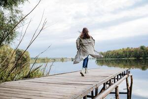 Rear view of a running girl in a raincoat on an old bridge on the lake, a woman from a run wants to jump into the water from the pier. photo