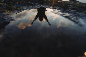 Reflection of the silhouette of a man in lying on a stone in the mountains, morning dawn, man raising his hands up, inverted image, sky reflected in water. photo