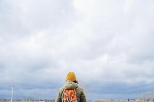 View from behind a person with a backpack on his shoulders and in a yellow hat, a tourist standing with his back against the sky, cloudy weather, clouds on foot, loneliness, silence. photo