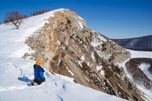 A little boy sits in the snow high in the mountains. A child travels through the winter snow climbing to the top of the mountain. Rocks against the blue sky. photo