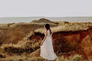 the bride walks on the field in the rays of the setting sun, the bride's white dress glows in the rays of the sun, the bride in a boho style hat, the concept of a wedding abroad photo