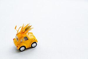 A small yellow car carries yellowed leaves. Autumn minimalistic composition with a toy car on a white background. photo