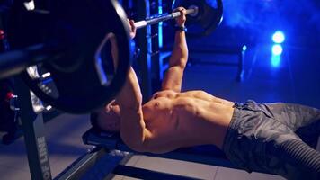 Sportsman training indoors. Young athlete lying on the board and lifting heavy barbell on the modern gym background. Blue light filter. video