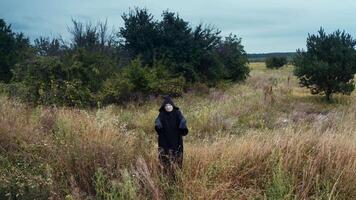 Scary death in black. Spooky figure in black mantle standing alone on the field near green trees. Drone view. Motion camera back. video