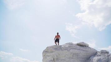 Sportive man on the edge of the hill. Shirtless man in shorts under the light sky. Muscular athlete on the top of mount in summer. View from below. Slow motion. video