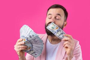 Portrait of a contented young businessman with a beard dressed in a pink jacket showing us dollar banknotes against a red studio background. Taste, smell of money. photo