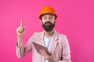 Portrait of young construction engineer wear orange hard hat, in a pink jacket standing on red studio background. A man with a tablet in his hands. photo