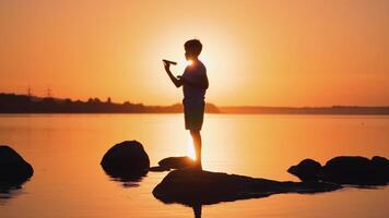 Silhouette of a boy with a paper plane at sunset. Child is standing on a stone on the orange river background. Bright rays of evening sun shines on a boy. video