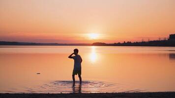 Backside view of a boy in the evening. Child standing in water and launching paper plane into the river at sunset. Slow motion. video