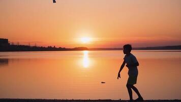 Boy near the river at sunset. Side view of a kid who is throwing paper plane up on the beautiful orange background of calm lake. Slow motion. video