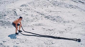Athletic male performs battle ropes exercise on the white rocky background. Sportsman in shorts doing waves with cable during an outdoor fitness workout. video