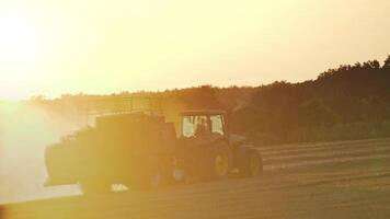 Tractor on the field during seasonal works at sunset. Agricultural machine pressing dry hay into square bale in the grassland on the trees background in the evening. video