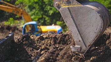 Bucket of tractor digs the ground. Excavator digging the soil on the background of green trees in summer. Close-up. video
