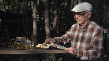 An elderly man sits at a wooden table, reading a book while sipping tea from a glass. Sunlight filters through the trees, creating a peaceful atmosphere in the woods. video