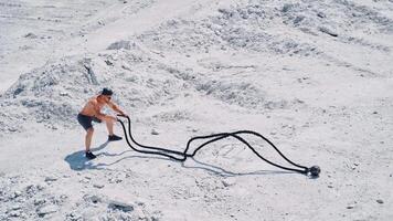 Strong young man doing exercises with a battle rope in sunny day. View from above on a muscular man training with cable on white ground outside. Concept of professional training. video