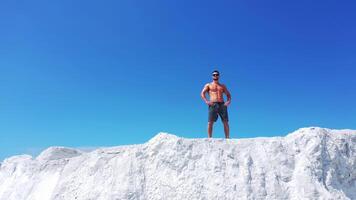 Drone view of a strong man in shorts without shirt on white hill. Young muscular man in good physical shape wearing sunglasses standing in nature. Camera moves up. video
