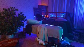 Worried, tired man sitting on bed at night and listening to music. Media. Young man can't sleep at night in a room with neon lighting. video