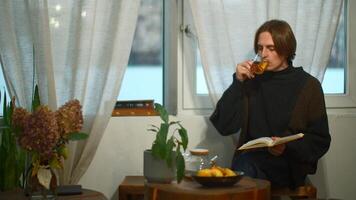 Young handsome man is reading book with tea in cafe. Media. Student is sitting alone and reading book in cozy cafe. Young man is reading book and drinking tea in student cafe video