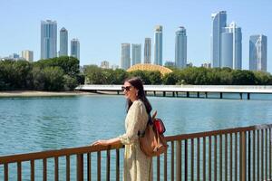 The skyline of Sharjah is visible from the waterfront - a woman tourist with a backpack enjoys the view of the water and skyscrapers. Cityscape of Sharjah UAE. photo