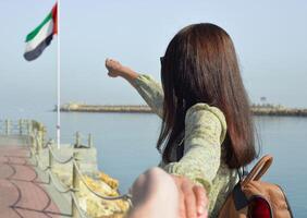 A young woman tourist in a long dress with a backpack points with her hand the direction to the sea to the flag of the United Arab Emirates, and leads her to the pier. photo
