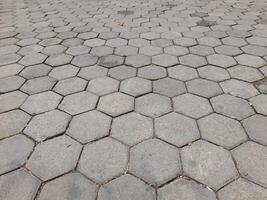 a pavement made of hexagonal concrete tiles arranged in a repeating pattern. The tiles are light gray and have visible wear, with some gaps and small debris between them. photo