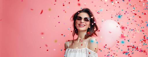 Portrait of young woman with sunglasses, surrounded by balloons and confetti on pink background photo